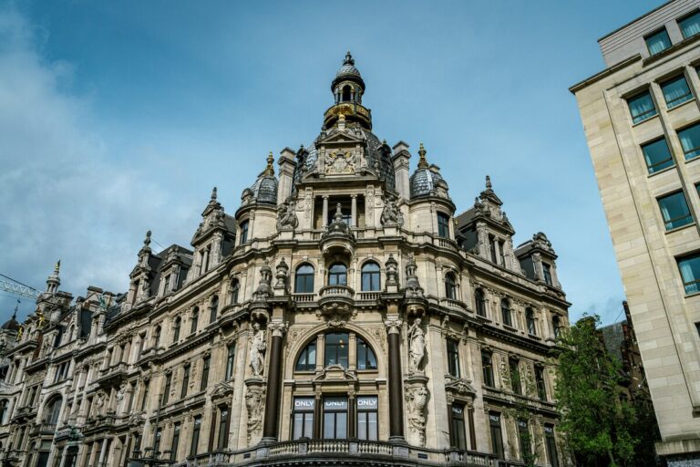 Façade ornée d’un bâtiment historique à Anvers, avec une architecture néoclassique et des détails sculpturaux raffinés. Le dôme central, richement décoré de dorures, domine l’édifice, entouré de statues et d’arcs élégants. Un exemple impressionnant du patrimoine architectural de la ville belge.