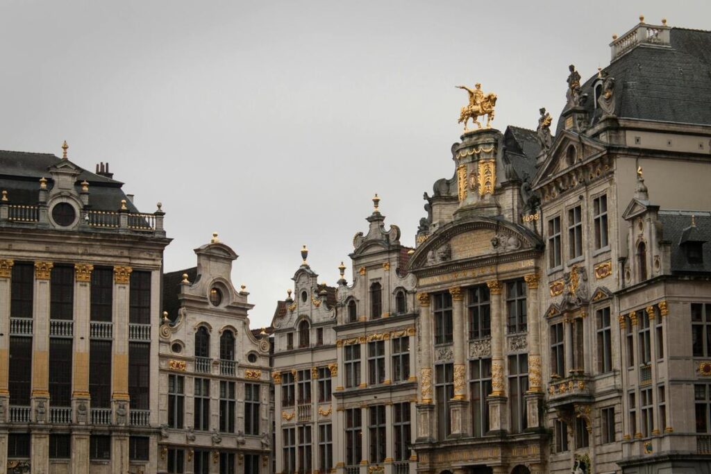 Détail architectural de la Grand-Place de Bruxelles, avec ses façades ornées de dorures, ses sculptures élaborées et son style baroque caractéristique. Les bâtiments historiques affichent des frontons décoratifs et des éléments néoclassiques, avec une statue dorée équestre dominant le sommet d'un édifice. Un symbole de l'héritage culturel et artistique de la capitale belge.