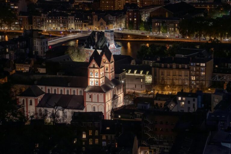 Vue nocturne de Liège, mettant en avant l'église Saint-Barthélemy illuminée, avec ses façades distinctives et son architecture romane. En arrière-plan, la Meuse traverse la ville, reflétant les lumières des ponts et des bâtiments environnants. Une scène captivante qui illustre le charme historique et urbain de Liège.