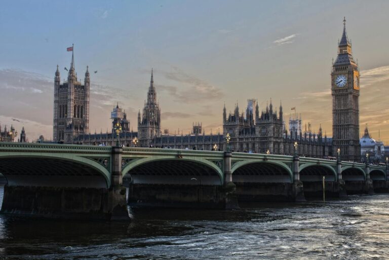 Vue emblématique de Londres au coucher du soleil, présentant le Palais de Westminster et Big Ben en arrière-plan. Au premier plan, le pont de Westminster traverse la Tamise, reflétant la lumière douce du ciel. L'architecture néogothique du Parlement se dresse majestueusement, soulignant l'histoire et la grandeur de la capitale britannique.