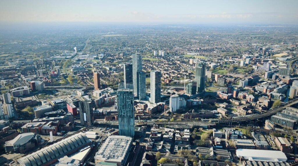 Vue aérienne de Manchester, mettant en avant ses gratte-ciels modernes, notamment les tours Beetham et Deansgate Square, entourés de bâtiments historiques et de quartiers résidentiels. La ville est traversée par des infrastructures ferroviaires et des routes principales, avec un panorama s'étendant sur les paysages verdoyants de la campagne anglaise à l'horizon. Une fusion entre urbanisme contemporain et patrimoine industriel.