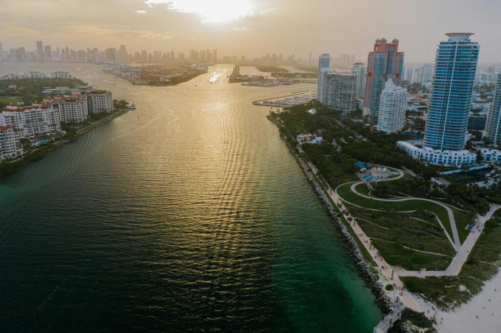 Vue aérienne de Miami au coucher du soleil, montrant la baie scintillante et les gratte-ciels modernes. À gauche, des résidences luxueuses bordent l'eau, tandis qu'à droite, un espace vert aménagé et des immeubles élégants bordent la côte. L'image met en valeur la beauté naturelle et urbaine de Miami, avec un contraste saisissant entre l'eau dorée et le ciel teinté par le soleil.