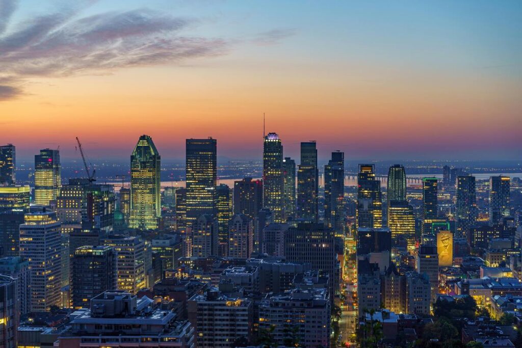 Vue panoramique de Montréal au crépuscule, mettant en valeur les gratte-ciels illuminés du centre-ville contre un ciel dégradé de couleurs chaudes. La silhouette emblématique de la ville est complétée par le pont Jacques-Cartier visible en arrière-plan et le fleuve Saint-Laurent reflétant les lumières de la métropole. Une scène urbaine dynamique et captivante.