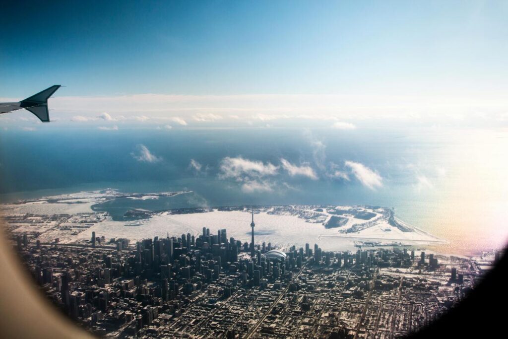 Vue aérienne hivernale de Toronto, capturée depuis un hublot d'avion, mettant en évidence la silhouette urbaine avec la Tour CN dominant les gratte-ciels. Le lac Ontario partiellement gelé s'étend à l'arrière-plan, créant un contraste saisissant avec le ciel bleu clair et les nuages épars. Une scène pittoresque illustrant la grandeur de la ville et la beauté naturelle environnante.