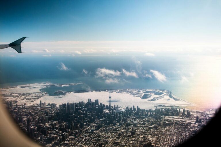 Vue aérienne hivernale de Toronto, capturée depuis un hublot d'avion, mettant en évidence la silhouette urbaine avec la Tour CN dominant les gratte-ciels. Le lac Ontario partiellement gelé s'étend à l'arrière-plan, créant un contraste saisissant avec le ciel bleu clair et les nuages épars. Une scène pittoresque illustrant la grandeur de la ville et la beauté naturelle environnante.