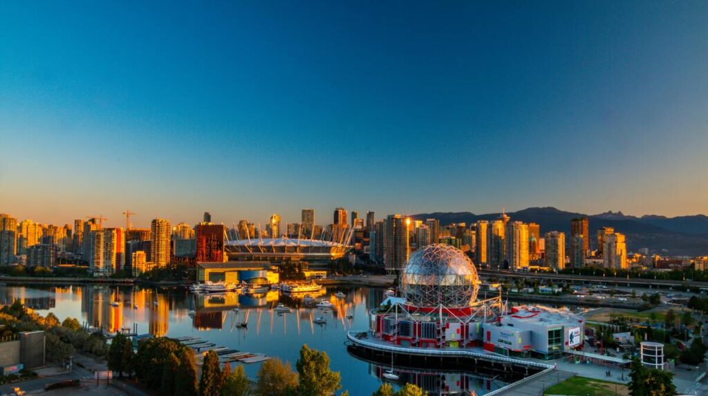 Vue panoramique de Vancouver au coucher du soleil, avec le Science World en premier plan et le stade BC Place illuminé à l'arrière-plan. Les gratte-ciels modernes se reflètent dans l'eau calme de False Creek, encadrés par les montagnes majestueuses à l'horizon. Une scène vibrante capturant l'équilibre entre nature et urbanisme.